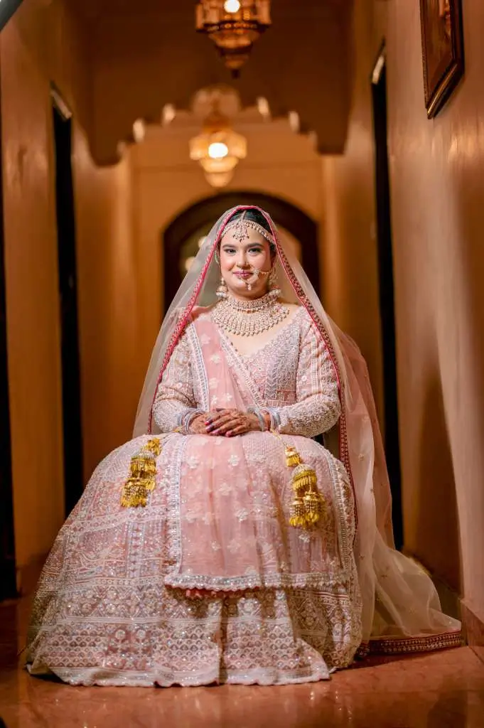 Elegant Indian bride seated gracefully in pastel pink wedding attire with traditional jewelry and warm indoor lighting.
