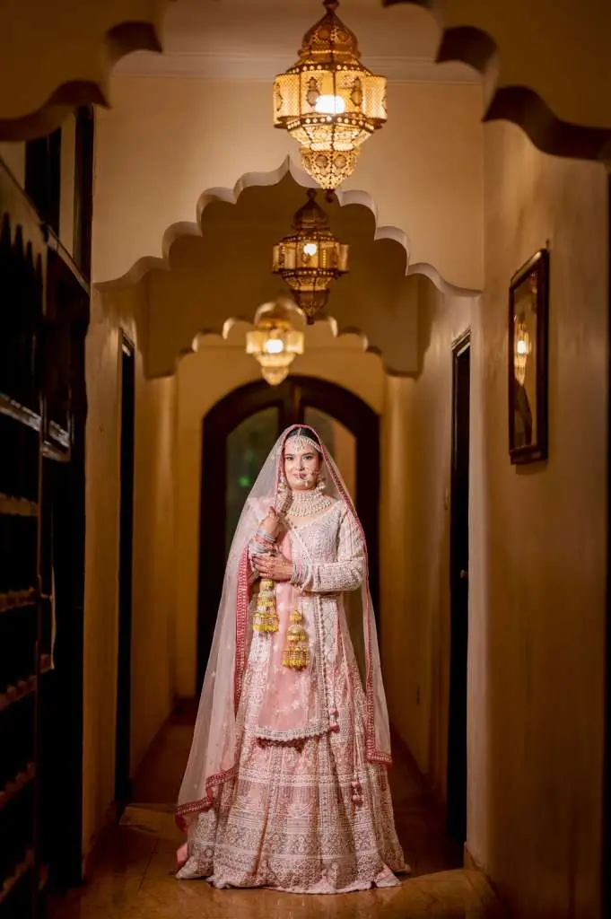 Full-length portrait of an Indian bride in pastel pink bridal attire standing in a royal hallway with decorative lantern lights.
