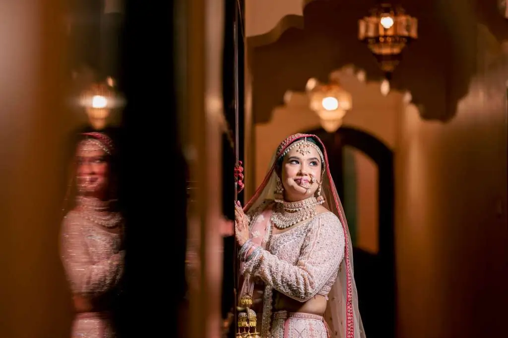 Candid portrait of an Indian bride in pastel pink bridal lehenga with artistic reflection effect in a luxurious indoor hallway.