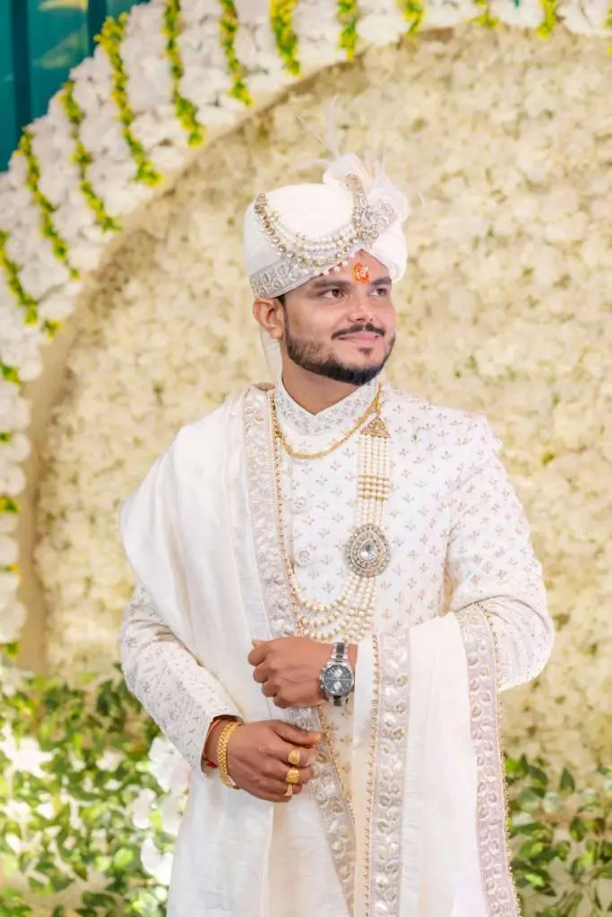 Elegant Indian groom portrait in traditional white sherwani and turban standing in front of a floral wedding backdrop.