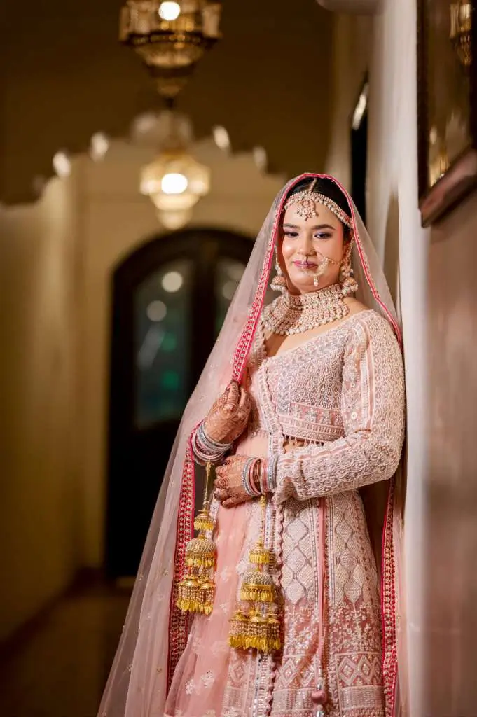 Royal style portrait of an Indian bride in pastel bridal lehenga standing in a decorative hallway with hanging lantern lights.